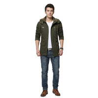 A man wearing an olive green hooded jacket with a brown faux-fur collar, styled over a white t-shirt and blue jeans against a light gray background.