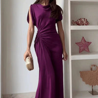 Woman wearing a long purple dress standing in a room with decorative items on shelves.