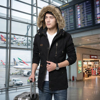 A man wearing a black hooded jacket with a tan faux-fur lined hood, front zipper, and flap pockets, paired with a white t-shirt and blue jeans at the airport.