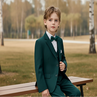 Young boy in a green suit with a bow tie sitting on a wooden chair against a plain background